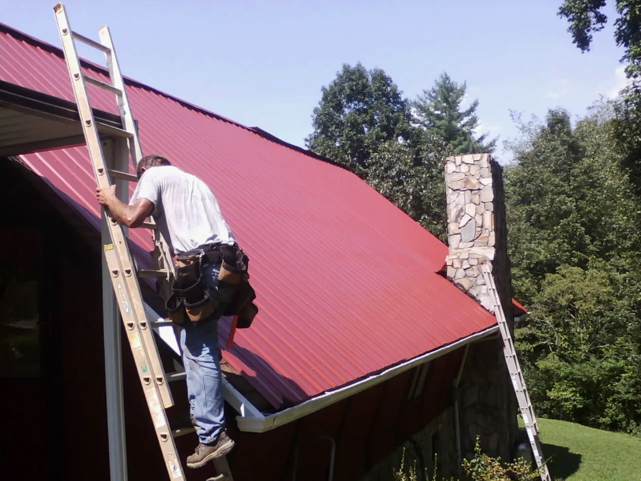 Roofer on a ladder beside a red metal roof and stone chimney — Soffit & Fascia Repair by Roofing Brothers in Creola
